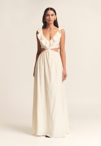 Largo vestido crema con un profundo escote en V, tirantes con volantes, cortes laterales y lunares negros, presentando una falda fluida y una cintura fruncida.