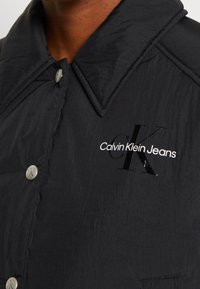 Sort nylonjakke med et glinsende Calvin Klein Jeans logo. Har en stor krave og knaplukning med metalaccenter. Glat tekstur.
