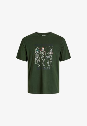 Adolescent portant un T-shirt vert foncé avec des squelettes et du texte, des manches longues blanches en dessous, un jean clair ample et des chaussures beiges, debout.