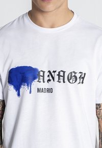 Biela bavlnená tričko s výrazným čiernym písmom a dizajnom modrého rozliatia farby. Text obsahuje "ANAGH" a "MADRID."