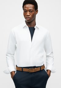 Άνδρας που φοράει λευκό πουκάμισο με κουμπιά πάνω από μαύρο τοπ, σκούρο παντελόνι και καφέ ζώνη, με τα χέρια στις τσέπες, μπροστά σε απλό φόντο.