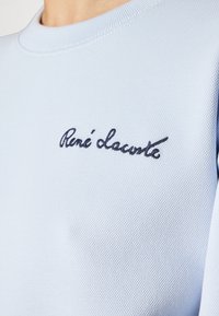 Chemise en tissu bleu clair texturé avec texte cursif bleu foncé « René Lacoste » brodé sur la partie supérieure de la poitrine.