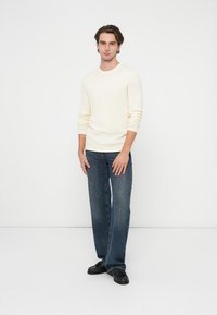 Jasnokremowy sweter z długim rękawem, proste ciemnoniebieskie dżinsy oraz czarne buty. Model stoi na neutralnym tle.