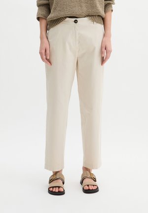 Beżowe spodnie o kroju 7/8 z zapięciem na guziki, noszone z jasnobrązowymi sandałami na klamrę oraz brązowym swetrem z dzianiny.