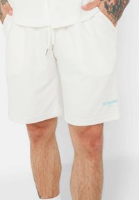 Shorts blancs en mélange de coton avec un design texturé, taille élastique et cordon de serrage. Présente le logo bleu "SIXTHUNE" sur la jambe gauche.