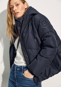 Veste rembourrée bleu marine avec capuche, à motif matelassé diagonal, présentant un écusson sur la manche et une fermeture éclair à l'avant. Associée à un jean bleu.