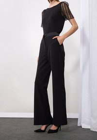 Černý outfit s přiléhavým topem se průhlednými rukávy posetými korálky a širokými kalhotami s vysokým pasem, doplněný černými lodičkami.