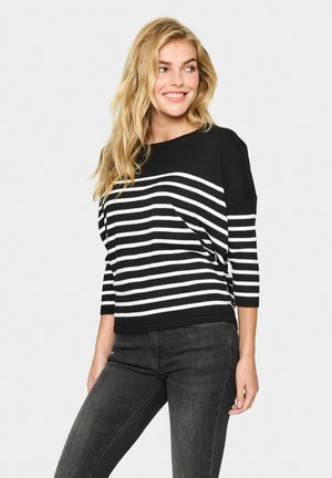 Pull en tricot noir et blanc à rayures avec un col bateau et des manches trois-quarts, associé à un jean foncé. Texture douce, design décontracté.
