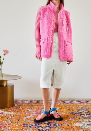 Femme portant un gilet polaire rose vif, un haut à manches longues rose côtelé, une jupe blanche à fines rayures avec une fente à l'avant, et des sandales colorées roses et bleues.