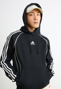 Jovem vestido com um hoodie preto da Adidas com riscas brancas e logo, chapéu bege, mãos nos bolsos do hoodie, em pé diante de um fundo branco plano.