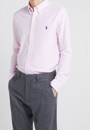 Man draagt een lichtroze gestreept overhemd met knopen en een blauw geborduurd logo en hoog getailleerde grijze broek met handen in de zakken.