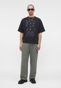 Mann trägt schwarzes oversized T-Shirt mit Totenkopf-Grafik, graue weit geschnittene Hose, schwarze Schuhe, Kettenkette und dunkle Sonnenbrille, steht mit entspannten Armen.