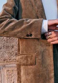 Brown corduroy blazer with vertical ribbing, single black button closure, and a front pocket. Underneath, a white shirt is visible.