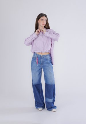 Mujer abrochándose una camisa corta de rayas lavanda, vistiendo jeans de pierna ancha bicolor y zapatos blancos, de pie contra un fondo claro y sencillo.