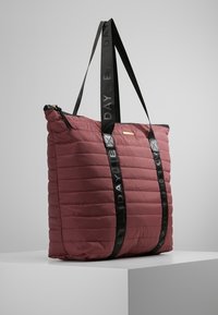 Bordeaux gekwilt handtasje met zwarte logo-handvaten, voorzien van een ritssluiting en glanzende accenten voor extra detail.