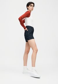 Νεαρός με κοντά σκούρα μαλλιά που φοράει γυαλιά, κόκκινο μακρυμάνικο μπλουζάκι κάτω από λευκό αμάνικο γιλέκο, σκούρες τζιν βερμούδες και λευκά ψηλά αθλητικά παπούτσια.