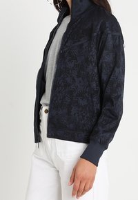 Námořnická bombardovací bunda s tonálním květinovým vzorem, zapínání na zip, žebrované manžety a límec, spárovaná se šedou košilí a bílými kalhotami.