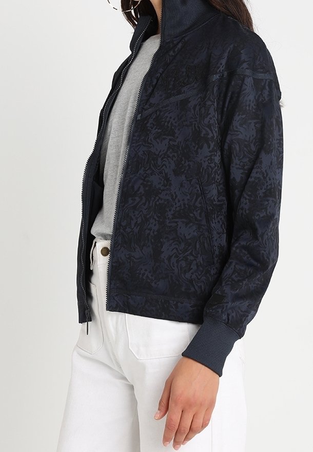 Námořnická bombardovací bunda s tonálním květinovým vzorem, zapínání na zip, žebrované manžety a límec, spárovaná se šedou košilí a bílými kalhotami.