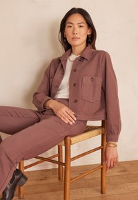 Giacca di jeans color ruggine con design aderente, tasca frontale e bottoni di legno. Abbinata a pantaloni coordinati e seduta su una sedia intrecciata.