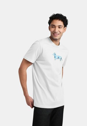 Giovane uomo sorridente, indossa una maglietta bianca con un motivo geometrico blu a forma di bassotto, in piedi con una mano dietro la schiena.