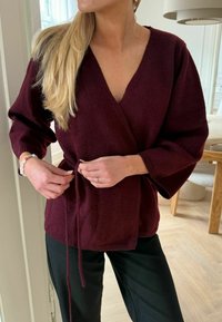 Pulover tricotat burgundy, cu guler în V și mâneci largi, prins cu un șnur. Purtați peste o pereche de pantaloni negri, cu un material texturat.