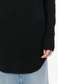 Pull noir en maille avec une texture côtelée, manches longues et un ourlet arrière plus long, porté sur un jean bleu clair.