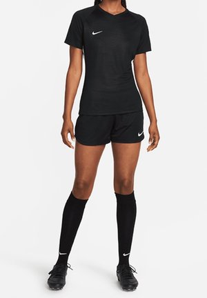 Kobieta stojąca, ubrana w czarny strój piłkarski Nike z krótkim rękawem, szortami, wysokimi skarpetami do kolan i korkami na białym tle.