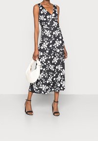 Svart blommig omslagsklänning med V-hals, prydd med vita blommönster, en midjlång kjol, kombinerad med en krämfärgad väska och svarta klackar med ankelsnörd.