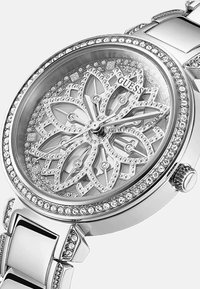 Zilveren horloge met een bloemmotief en kristallen versieringen, en een gepolijste metalen band. De wijzerplaat is doorschijnend met een gestructureerd patroon.