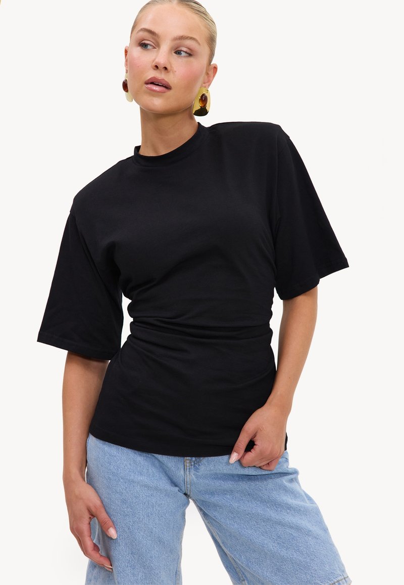 Femme portant un haut ajusté à manches courtes noir et un jean bleu clair, avec des boucles d'oreilles pendantes en or et ambre, posant avec une main sur la hanche.