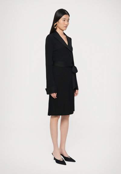 Μαύρο φόρεμα τύπου wrap με μακριά μανίκια και ζώνη στη μέση. Διαθέτει βαθύ V-λαιμόκοψη και μανσέτες με κουμπιά. Φορεμένο με μαύρες γόβες μυτερές.