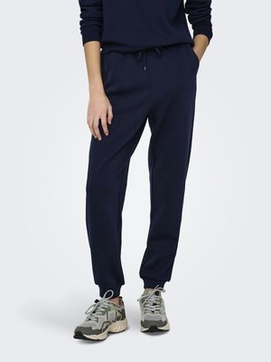 Personne portant un pantalon de jogging bleu marine et des baskets à motif camouflage, debout les mains dans les poches devant un fond uni.
