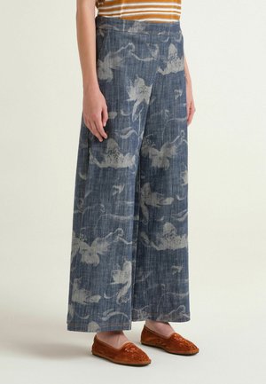 Frau trägt weite Jeanshosen mit weißen Blumenmustern und braunen bestickten Slippern, stehend vor einem hellen Hintergrund.