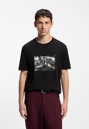 Jeune homme portant un T-shirt noir avec une impression tableau de bord de voiture en noir et blanc et un pantalon bordeaux, debout devant un fond blanc uni.