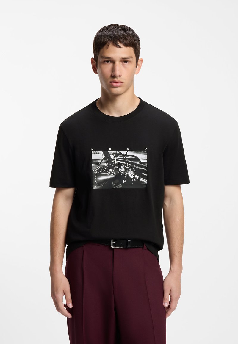 Jeune homme portant un T-shirt noir avec une impression tableau de bord de voiture en noir et blanc et un pantalon bordeaux, debout devant un fond blanc uni.