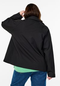 Giacca nera oversize con colletto strutturato, maniche lunghe e una texture liscia, indossata sopra una camicia verde e jeans blu.