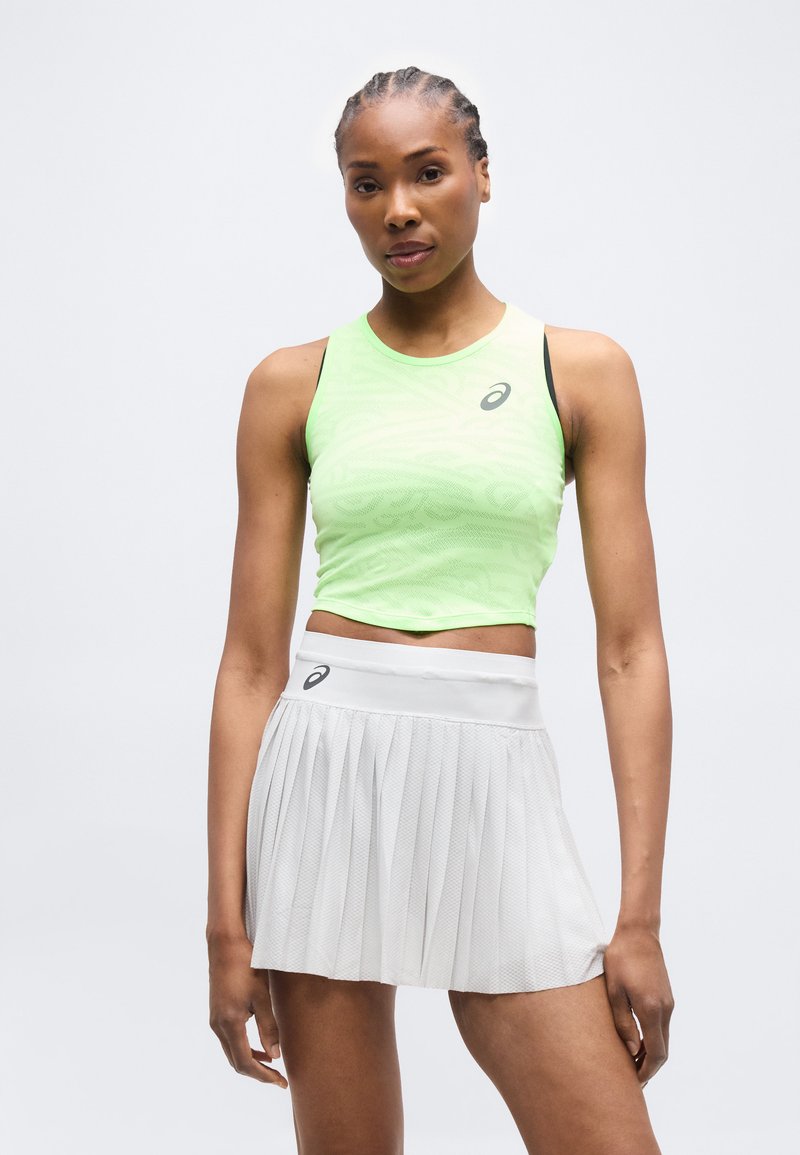 Αθλητική γυναίκα που φοράει ένα ανοιχτό πράσινο αμάνικο κορυφαίο τοπ και λευκή πλισέ αθλητική φούστα, στέκεται μπροστά σε ένα απλό φόντο.