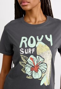 T-shirt gris avec un design graphique comprenant le texte "ROXY SURF", un poisson et une grande fleur d'hibiscus avec des accents vert clair et rouge.