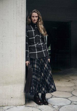 Schwarz-weiß kariertes Hemd, langer karierten Rock in dunklen Tönen, braune Stiefeletten, hält eine strukturierte dunkle Tasche, steht neben einer Betonwand.