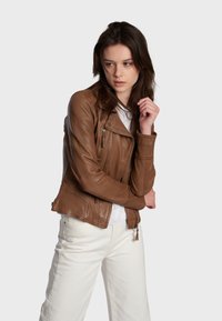 Braune Leder-Motorradjacke mit Reißverschlusstaschen, strukturierter Oberfläche und asymmetrischem Frontverschluss, kombiniert mit weißen Hosen.