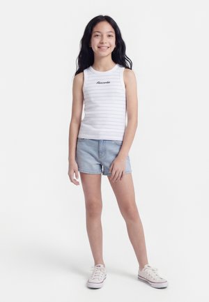 Νεαρή κοπέλα που στέκεται και χαμογελά φορώντας λευκό ριγωτό αμάνικο τοπ, ανοιχτόχρωμο τζιν σορτς και λευκά αθλητικά παπούτσια σε λευκό φόντο.