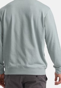 Hombre vestido con un suéter de manga larga gris claro y pantalones gris oscuro, visto desde atrás con las manos relajadas a los lados.