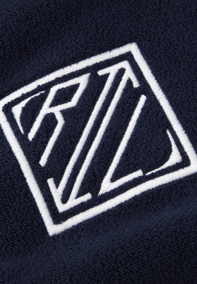 Gros plan sur un tissu bleu marine avec un logo géométrique brodé en blanc, présentant les lettres stylisées R et L à l'intérieur d'une forme losange.