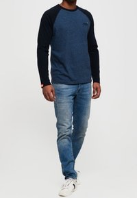 Katoenen blouse met lange mouwen met een blauwe romp en zwarte raglanmouwen, gecombineerd met lichtblauwe jeans en witte sneakers op een neutrale achtergrond.