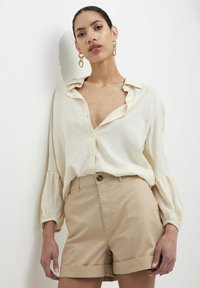 Camicia beige a maniche lunghe con vestibilità rilassata e chiusura a bottoni, abbinata a pantaloni corti di colore beige chiaro con orli arrotolati. Modello con orecchini statement.