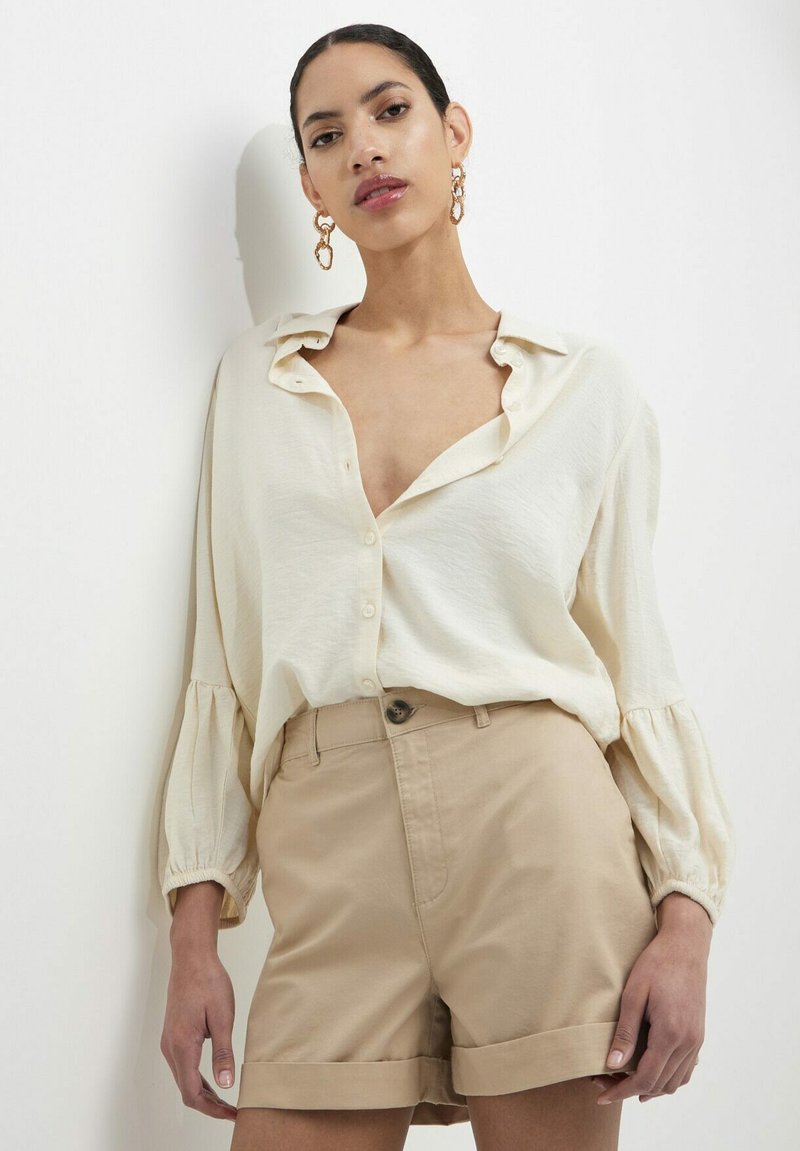 Camicia beige a maniche lunghe con vestibilità rilassata e chiusura a bottoni, abbinata a pantaloni corti di colore beige chiaro con orli arrotolati. Modello con orecchini statement.