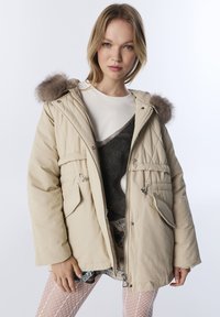 Beige gewatteerde parka met capuchon, voorzien van een afneembare bontkraag, voorzakken en een taille met trekkoord. Gladde textuur, casual ontwerp.