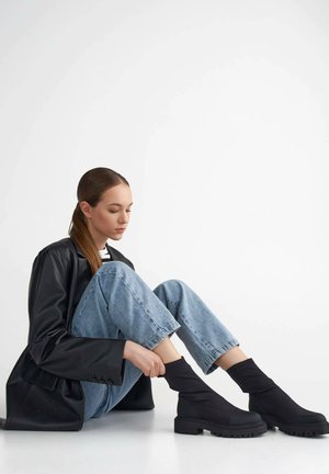 Black ankle boots made of textured fabric, featuring a high sock-like shaft and a chunky rubber sole with defined tread. Worn with blue jeans.