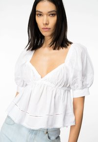 Blusa in cotone bianco con maniche a sbuffo, scollo a V profondo e vita arricciata. Presenta trame di pizzo e dettagli a taglio lungo l'orlo. Modella che indossa jeans blu.