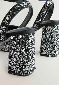 Black high-heeled sandals with chunky block heels covered in various white and silver sequins and beads, featuring satin straps.